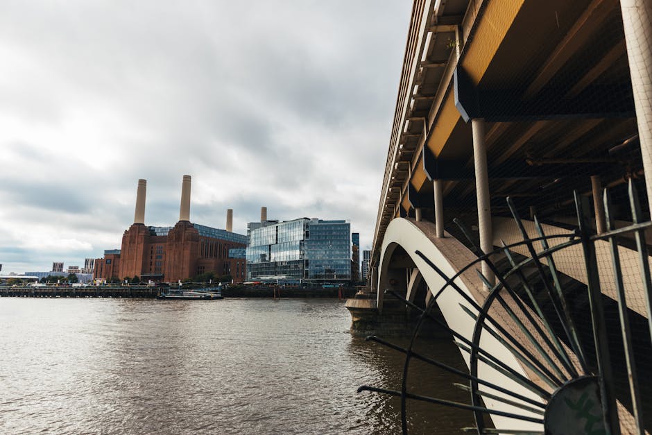 A view of the river Thames in Battersea during overcast weather, with the iconic Tate Modern building featuring its red brick exterior and four chimney stacks visible in the background on the left. In the foreground on the right, part of a modern bridge structure with metal supports and a white, curved architectural element is seen, extending beneath the bridge deck. The scene captures a mix of industrial and contemporary architecture, with the water reflecting the cloudy sky above. This setting aligns with the context of rubbish removal or waste clearance by highlighting urban infrastructure near residential or commercial areas, where independent waste collection services such as those offered by Waste Clearance Battersea might operate, especially in relation to property or site clearance projects near Battersea's iconic landmarks.