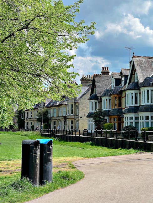 A residential street scene featuring a row of Victorian-style terraced houses with bay windows, slate roofs, and brick chimneys, situated behind a black wooden fence. In the foreground, there are two black cylindrical rubbish bins placed on a grassy verge near a concrete footpath. To the left, a large leafy tree with bright green foliage extends its branches over the scene, contrasting with the partly cloudy sky overhead. The environment appears clean and well-maintained, typical of an urban neighbourhood where local waste management services or private rubbish collection might be utilized, aligning with the context of private waste handling and alternative rubbish removal options mentioned on the page.