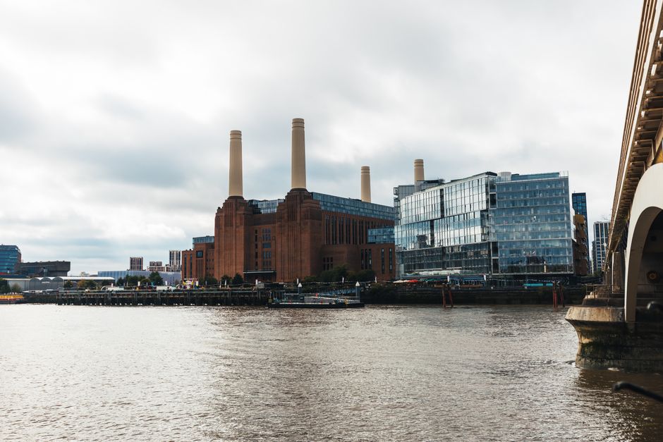 A large industrial building with a red brick exterior and four tall, beige smokestacks dominates the scene, situated beside a river. The structure features rectangular windows and a solid, utilitarian design characteristic of a power station or manufacturing facility. To the right of the industrial complex, a modern glass-fronted office building with reflective windows is visible, providing a contrast in architectural styles. The river in the foreground has a calm, rippled surface with a brownish hue, likely reflecting the cloudy sky overhead. Part of a stone and metal bridge is visible on the right side of the image, with the stone pier supporting its structure. The environment appears to be an urban riverside area with some industrial and contemporary elements, set under an overcast sky that creates diffuse, soft lighting. This scene relates to waste management and rubbish removal services, capturing the industrial and urban landscape often associated with large-scale rubbish clearance and private waste handling operations by companies such as Waste Clearance Battersea.