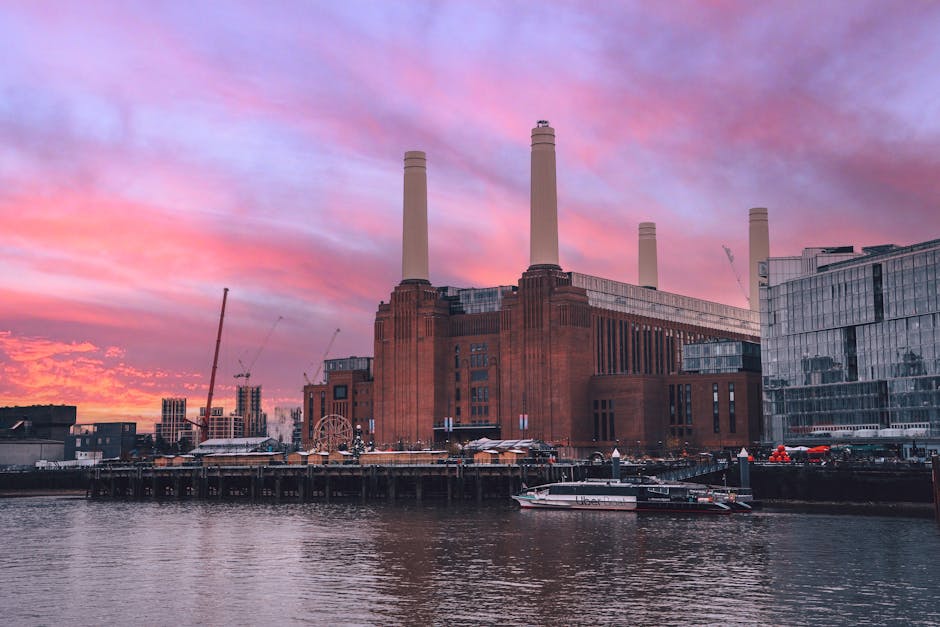 A large, historic power station built with red brick, featuring four tall, white industrial chimneys extending upward against a colorful sunset sky with shades of pink, purple, and orange. In the foreground, there is a body of water with a white and black speedboat docked along the pier. Surrounding the power station are modern glass-fronted buildings reflecting the sky's hues, and construction cranes can be seen in the background, indicating ongoing development. The scene captures the mix of industrial and modern architecture typical of a waterfront area near Battersea. The environment appears calm, with the water gently rippling and the sky providing a vibrant backdrop, subtly emphasizing the context of urban regeneration and alternative waste handling practices in the vicinity, which aligns with services of rubbish removal and private disposal providers like Waste Clearance Battersea.