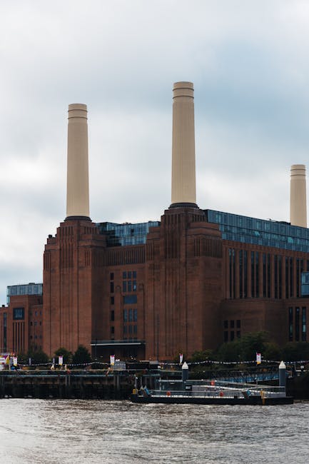A large industrial building with a red brick exterior and four tall, beige smokestacks dominates the scene, situated beside a river. The structure features rectangular windows and a solid, utilitarian design characteristic of a power station or manufacturing facility. To the right of the industrial complex, a modern glass-fronted office building with reflective windows is visible, providing a contrast in architectural styles. The river in the foreground has a calm, rippled surface with a brownish hue, likely reflecting the cloudy sky overhead. Part of a stone and metal bridge is visible on the right side of the image, with the stone pier supporting its structure. The environment appears to be an urban riverside area with some industrial and contemporary elements, set under an overcast sky that creates diffuse, soft lighting. This scene relates to waste management and rubbish removal services, capturing the industrial and urban landscape often associated with large-scale rubbish clearance and private waste handling operations by companies such as Waste Clearance Battersea.