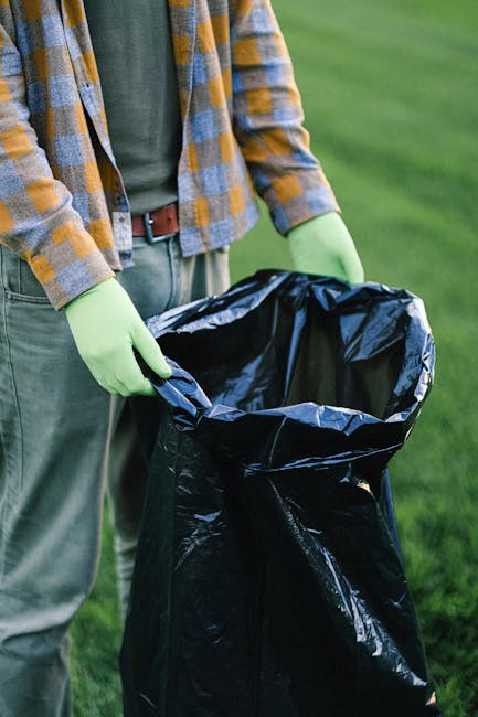 The image shows a person wearing a yellow and gray checked shirt over a dark green T-shirt, standing outdoors on a grassy area. The individual is wearing light green disposable gloves and holding a large black rubbish bag, which is partially open. The bag appears to be made of plastic with a glossy finish, and it is situated in front of the person's waist, with the top edges folded outward. In the background, there is a well-maintained, lush green lawn, indicating an outdoor environment suitable for waste collection or rubbish clearance activities. The scene suggests a context of manual rubbish removal, possibly involving private waste handling or community clean-up, and it aligns with services such as those offered by Waste Clearance Battersea for efficient rubbish disposal.