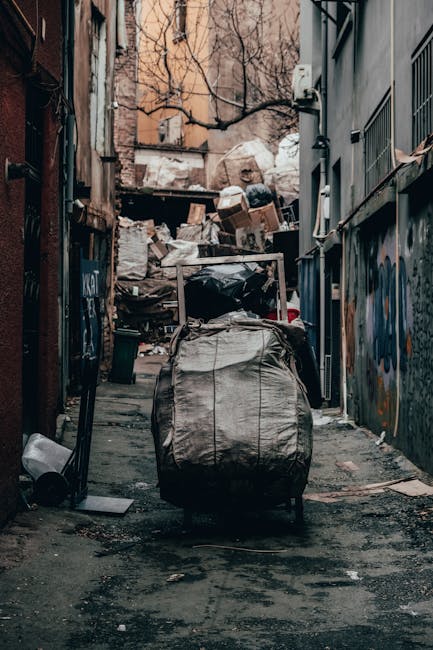 A narrow urban alleyway with a concrete surface scattered with small debris and dirt. At the center, a large, grey, weathered garbage bag with visible creases and stains is placed on a trolley or cart, which appears to be used for rubbish collection. The alley is enclosed by tall, multi-storey buildings on both sides, featuring graffiti on the right wall and an array of utility panels, electrical boxes, and pipes. In the background, an accumulation of mixed waste, including cardboard boxes, plastic bags, and other refuse, is piled up against a wall, partially covered with a large white sack or tarp. Leafless tree branches are visible overhead, suggesting an outdoor setting during colder months. The scene has a dim, natural light atmosphere, typical of alleyways used for waste disposal, aligning with private or alternative rubbish removal practices, as managed by waste services like Waste Clearance Battersea.